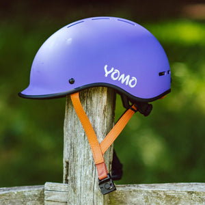 YOMO matt purple kids' helmet perched on a wooden post against a blurred, natural background.
