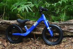 HOY Weee! blue balance bike resting against a log, with green foliage in the background.
