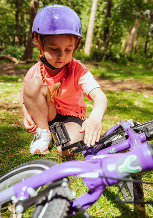 Child wearing a YOMO glossy purple kids' helmet and sitting on the grass, next to a YOMO purple bicycle.