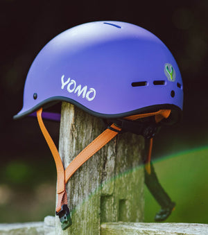 YOMO matt purple kids' helmet perched on a wooden post against a blurred, natural background.