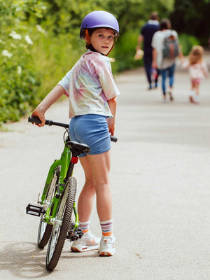 Child wearing a YOMO matt purple kids' helmet with a YOMO 20 green bike with people in the background.
