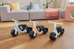 Three Woom WOW balance bikes on a wooden floor with a sofa and cushions in the background.